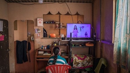 A peruvian student sits in a red plastic chair watching an educational broadcast at home while unable to attend school due to the COVID-19 pandemic. 